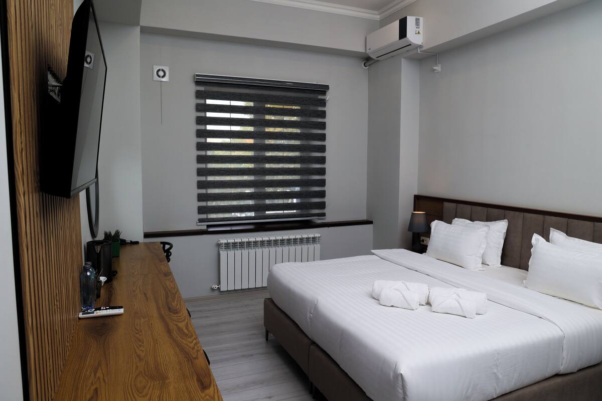 A modern hotel room with a large bed, a wooden dresser, a flat-screen TV, and a window with blinds.