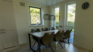 A modern dining area with a wooden table and six chairs, large windows with a view of greenery, and a wall clock. - Holiday home in Ewijk in a beautiful area (Ewijk)