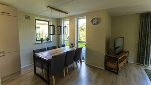 Une cuisine moderne avec une table à manger en bois, des chaises, des luminaires suspendus, un téléviseur encastré et une grande fenêtre donnant sur un paysage verdoyant.