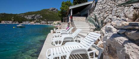 Un hôtel en bord de mer, doté d’une façade en pierre, d’une eau d’un bleu limpide et de chaises longues blanches au bord de la piscine.