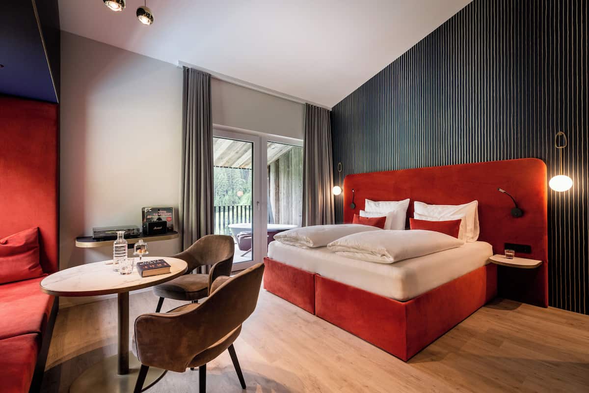 A modern hotel room with a red bed, a round table, a chair, and a view through a glass door.