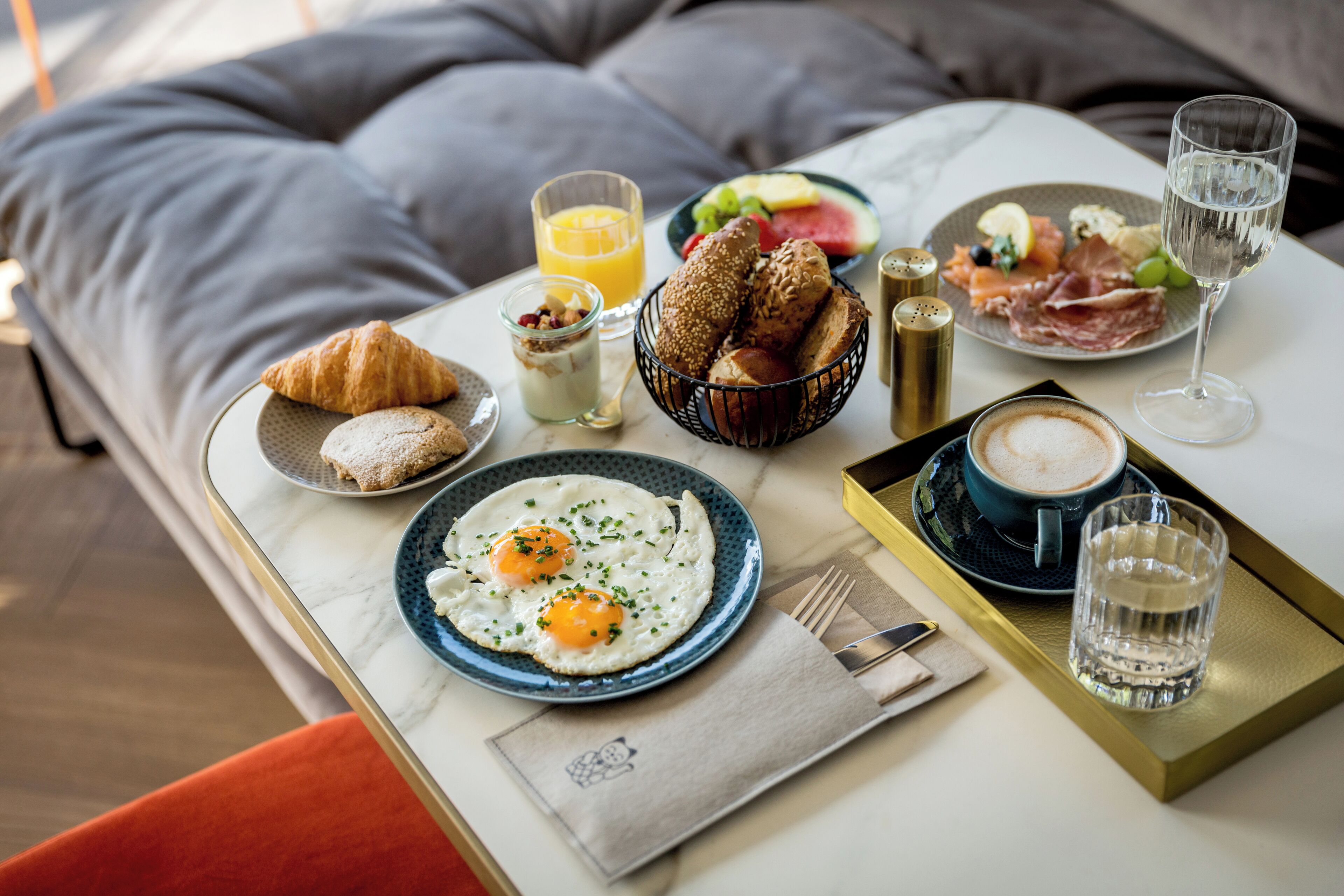 Una mesa con desayuno que incluye cruasanes, huevos, tostadas, fruta y bebidas.