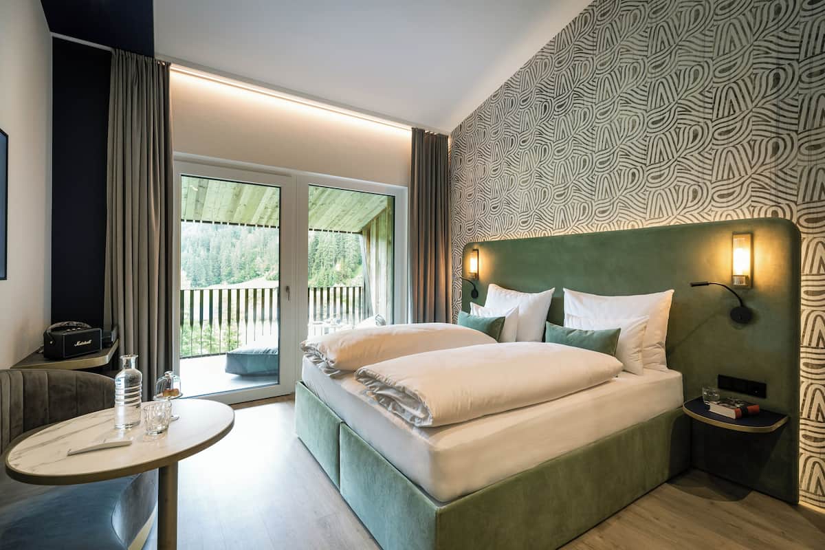 A modern hotel room with a large bed, a small table, a sofa, and a balcony with a view of greenery.