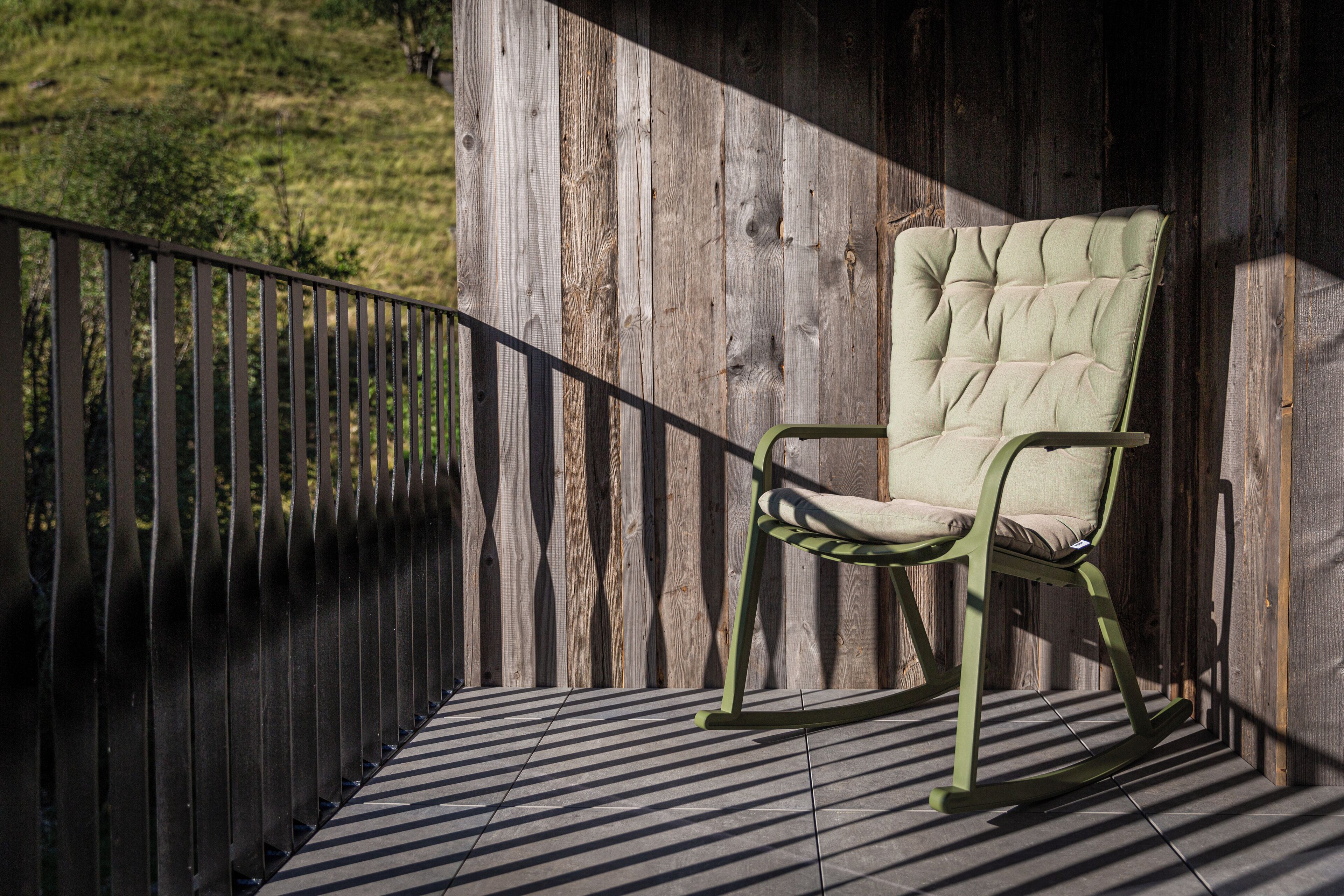 A rocking chair on a wooden deck with a metal railing, set against a wooden wall.