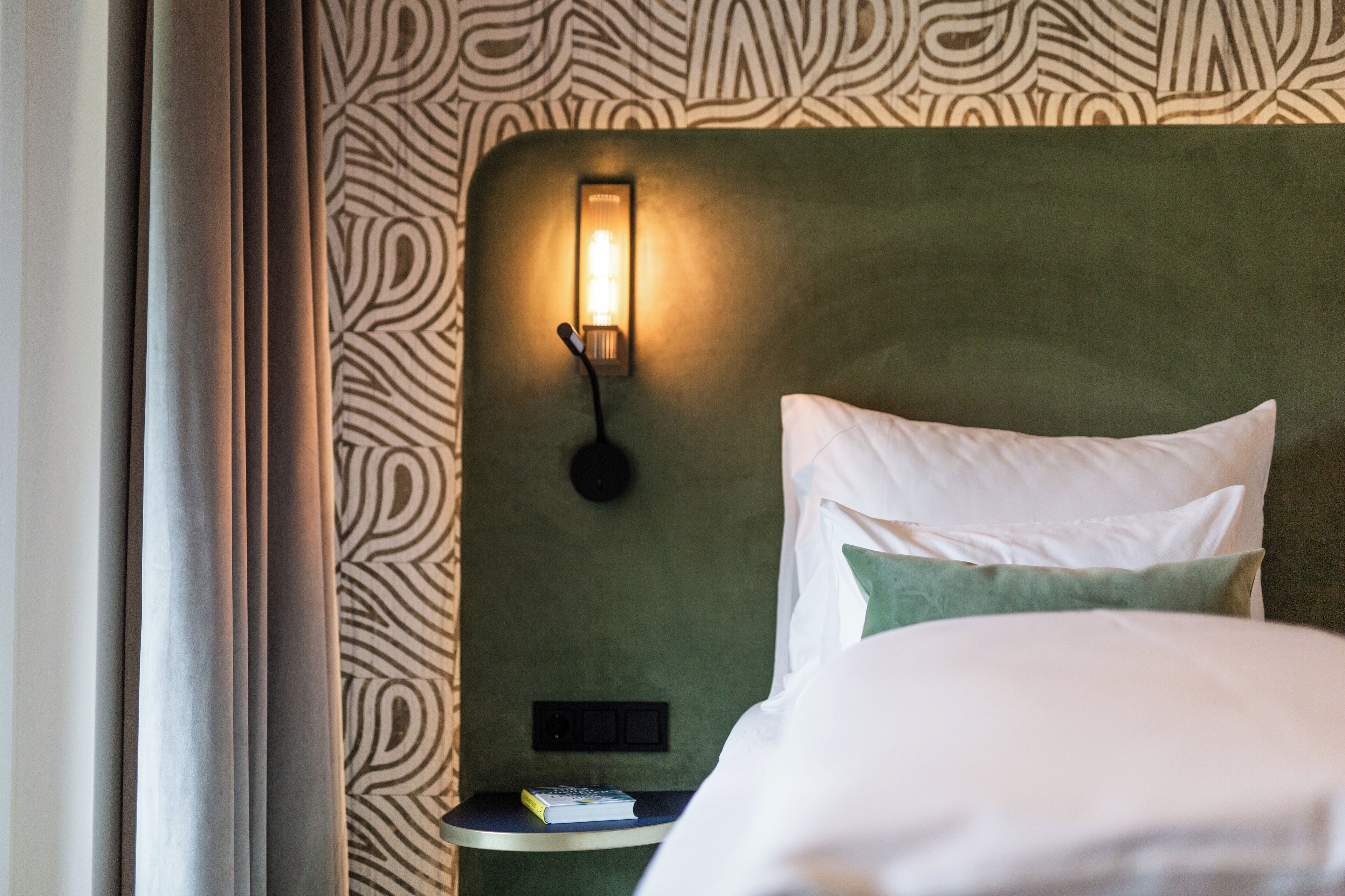 A neatly made bed with white and green pillows, a bedside table with a book, and a wall-mounted light fixture.
