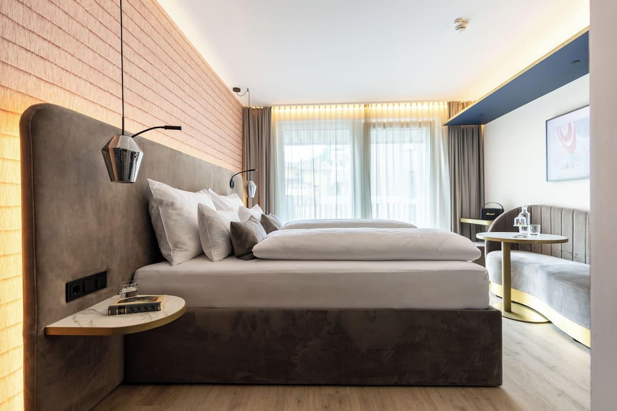A modern hotel room with a large bed, a bedside table with a lamp, a small round table, and a view of the city through large windows.