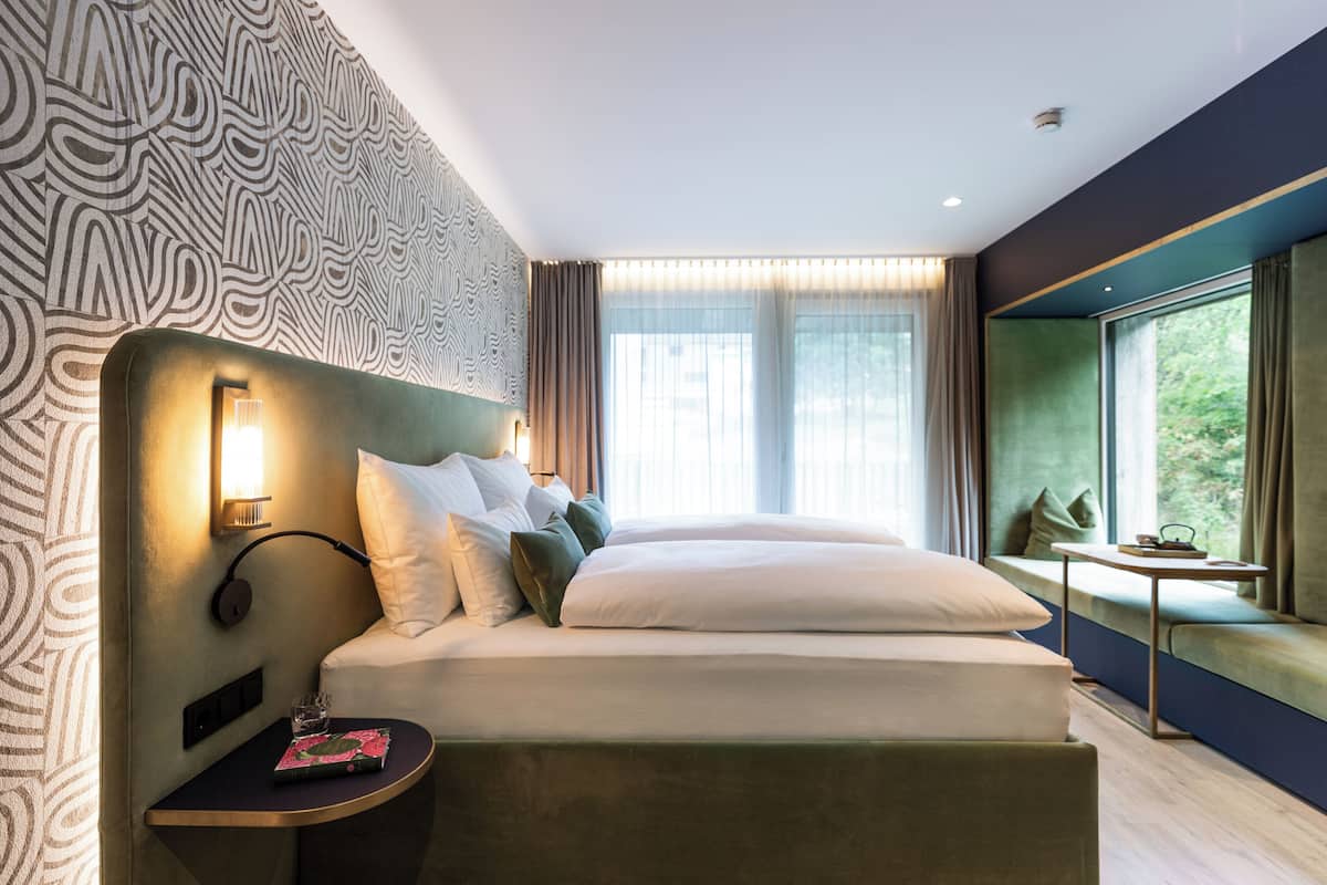 A modern hotel room with a large bed, a bedside table with a lamp, a window seat, and a patterned wall.