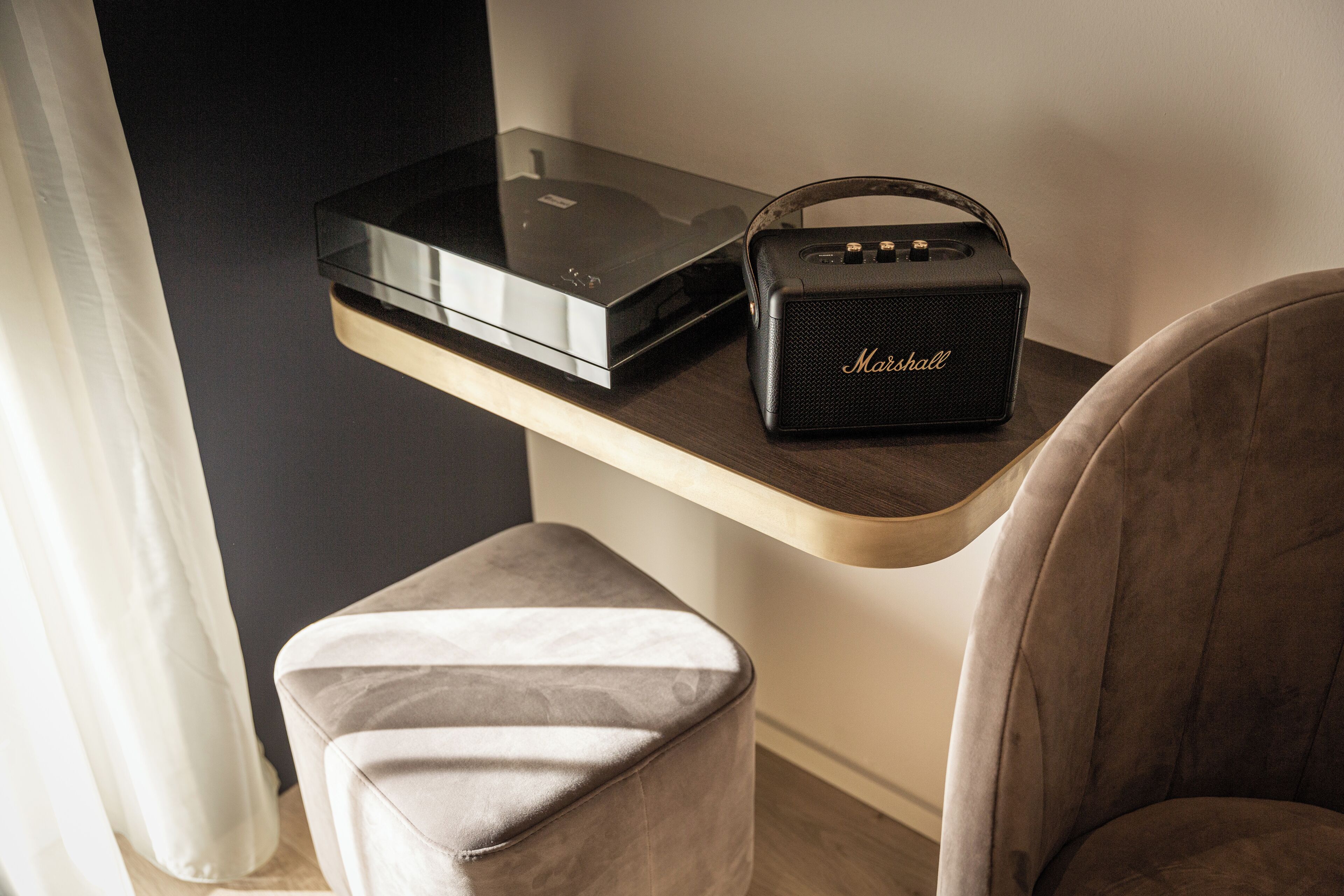 A modern room with a flat-screen TV on a wooden shelf, a Marshall speaker, and a cushioned ottoman.