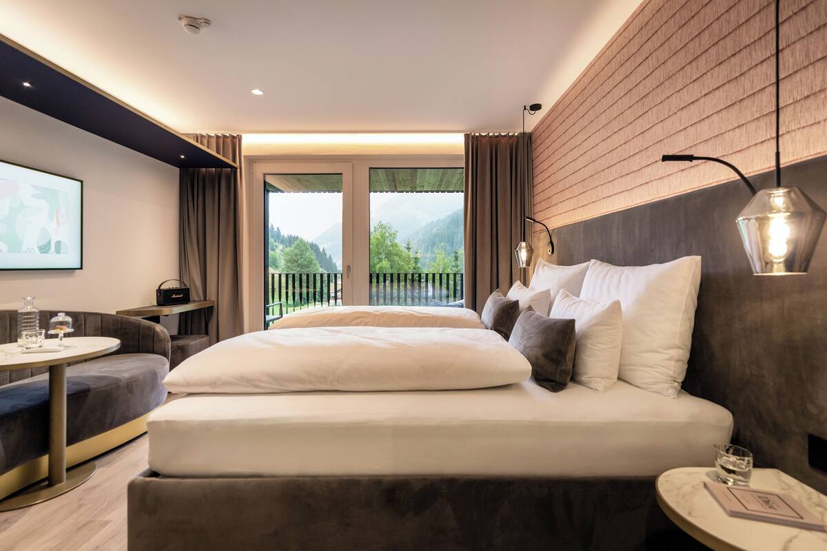 A modern hotel room with a large bed, a seating area with a table, and a balcony with a view of greenery and mountains.