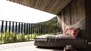 Un balcon offrant une vue sur une chaîne de montagnes boisées, un coin salon confortable avec des coussins et un oreiller, ainsi qu’une table avec une tasse et des livres.