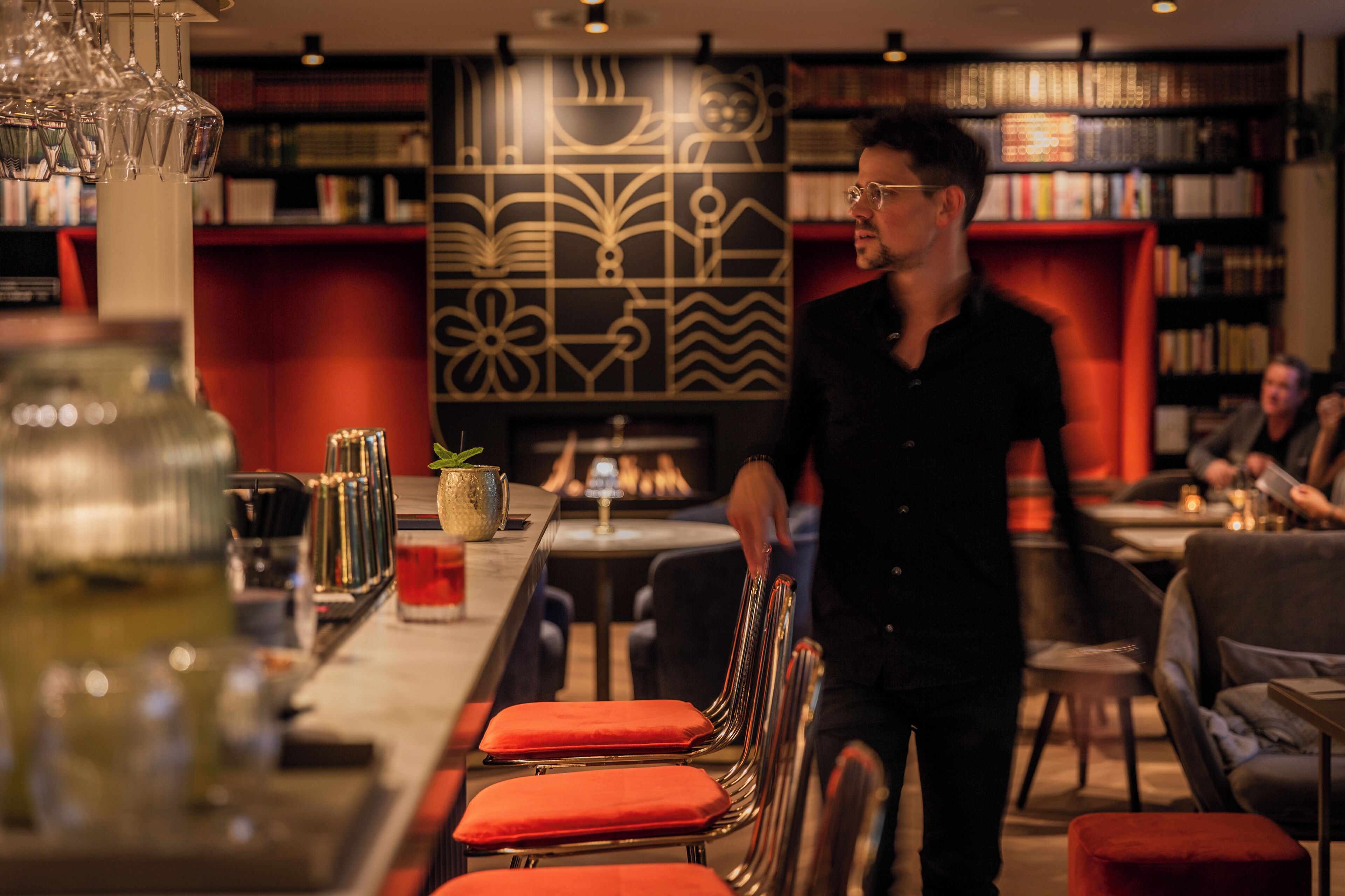 Una zona de bar con mostrador, taburetes con cojines rojos y una pared decorada al fondo.