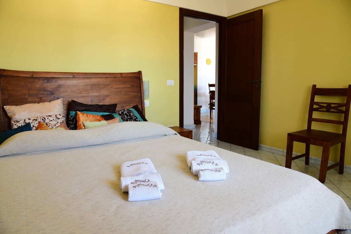 Un letto rifatto con cura, una testiera in legno, due asciugamani piegati e una selezione di cuscini.