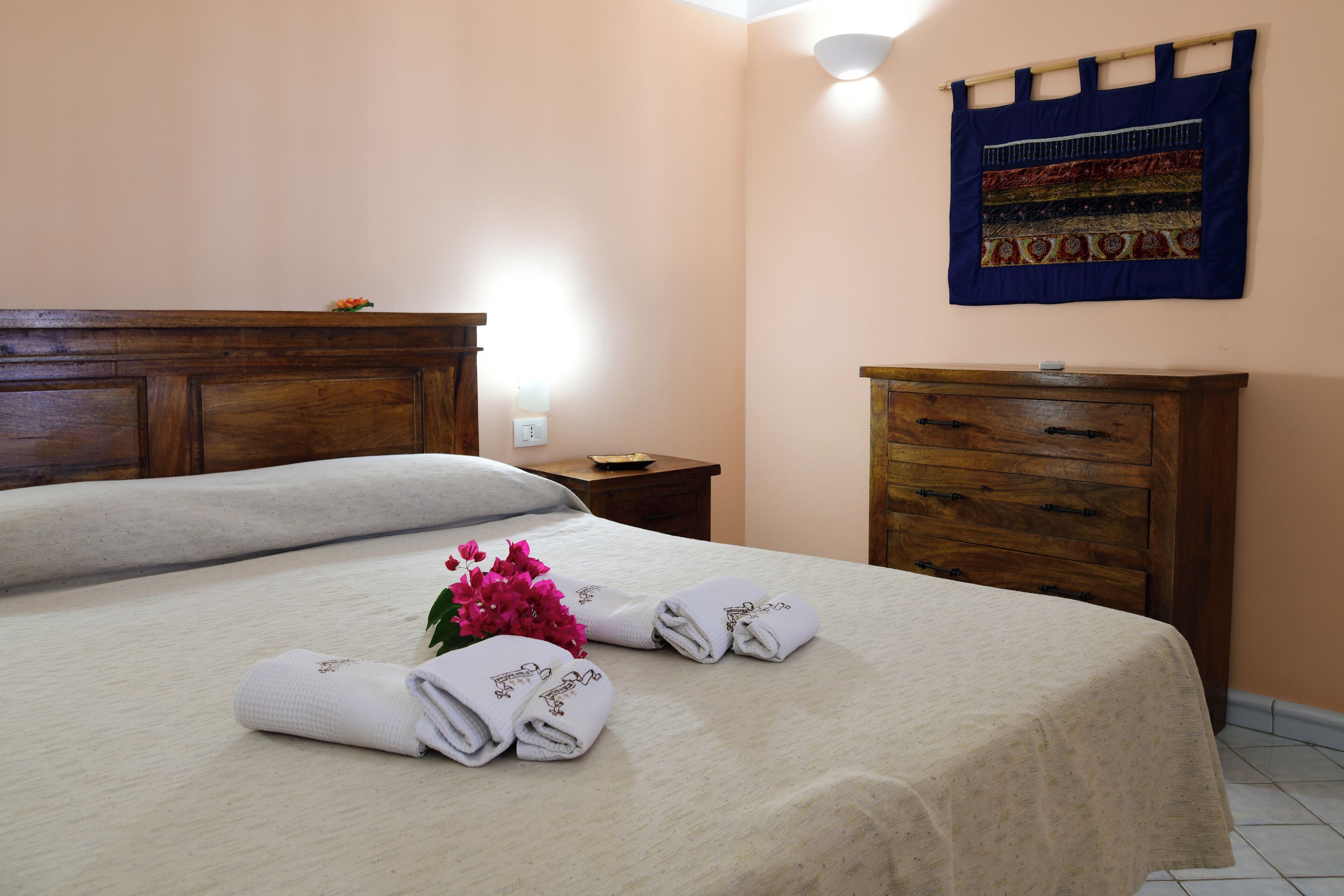 Una cama bien tendida con toallas dobladas y un jarrón de flores, un aparador de madera y un tapiz colgado en la pared.