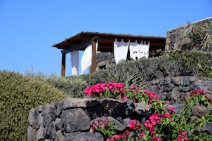 A stone structure with a wooden roof and white curtains, surrounded by greenery and pink flowers.
