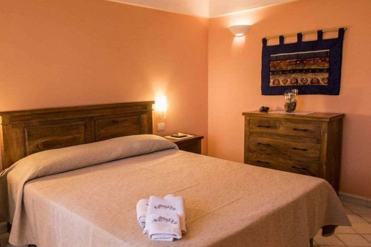 Un letto rifatto con copripiumino color beige e cuscini, un comodino in legno con una lampada, una cassettiera in legno con uno specchio e un tappezzeria a parete.