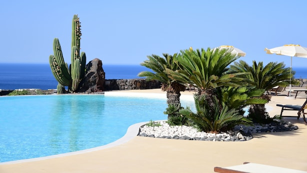A pool with clear blue water, surrounded by rocks and cacti, with a view of the ocean and beach chairs. - Dammusi Sotto le Stelle (Pantelleria)