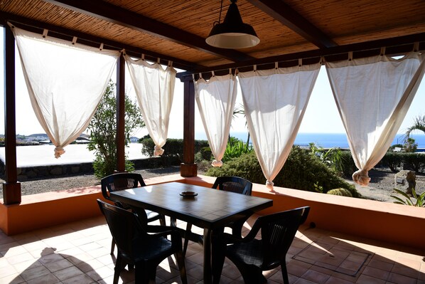 Un patio cubierto con una mesa y sillas de madera, cortinas blancas y vista al mar.