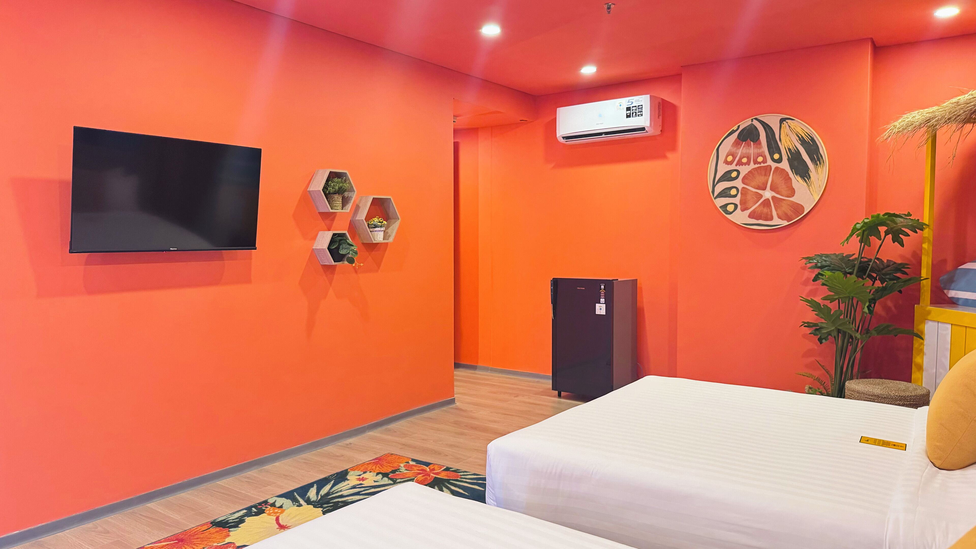 A hotel room with orange walls, a flat-screen TV, a refrigerator, and a bed with white bedding.