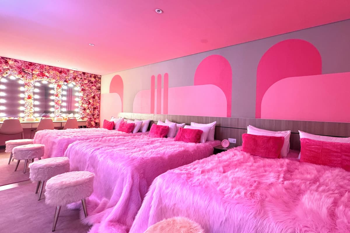Habitación de hotel con ropa de cama rosa, un tocador con espejos y una decoración floral en la pared.