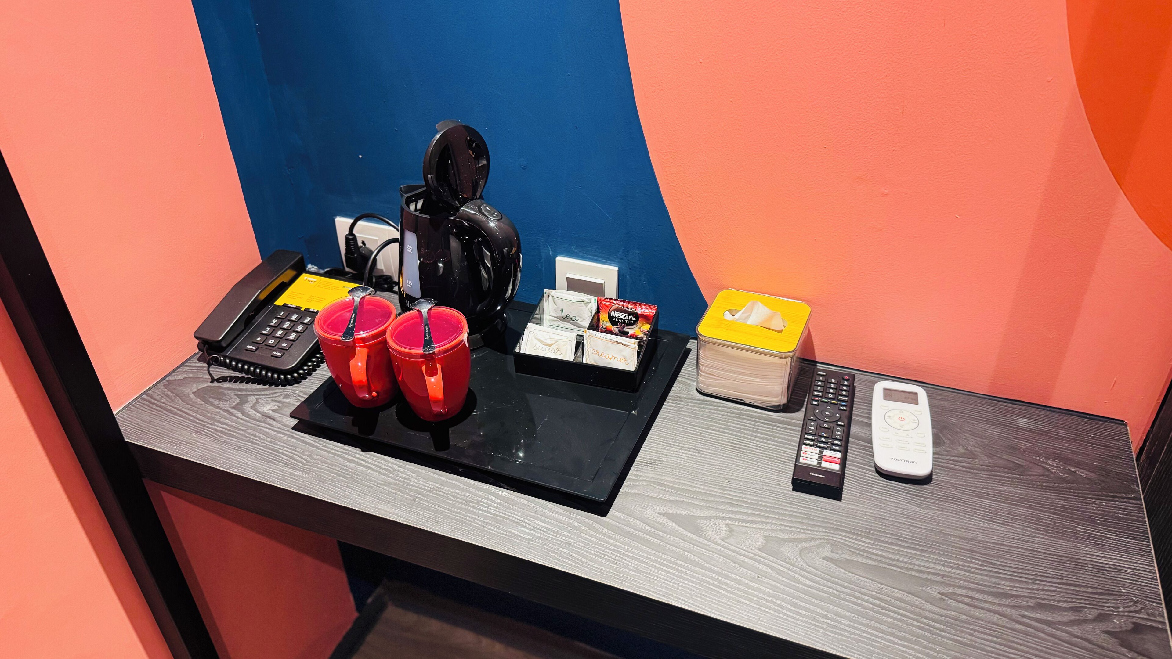 En hotellreception med en telefon, en kaffemaskin, två röda muggar, en näsdukspåse och en fjärrkontroll.