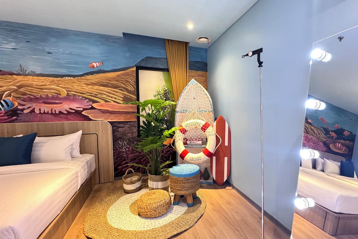 Una habitación con un mural de temática náutica, una cama, un pequeño rincón de descanso con una tabla de surf y un espejo con luces.