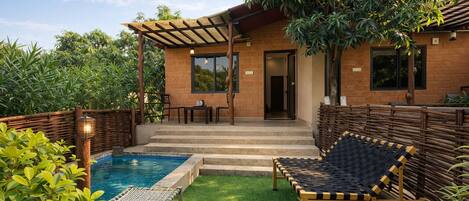 A poolside area with a pool, two lounge chairs, a pergola, and a brick building with a door.