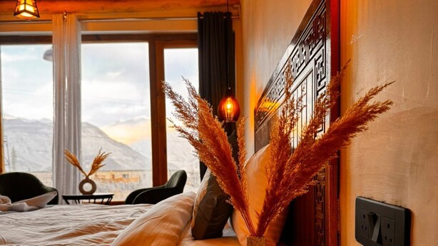 Ein Schlafzimmer mit einem Bett, einem Nachttisch aus Holz, einer Vase mit Trockenpflanzen, einer Wandleuchte und Blick auf Berge durch ein Fenster.