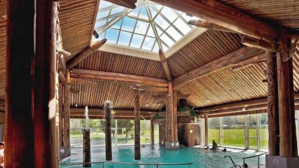 A large indoor swimming pool with a wooden roof and glass ceiling, surrounded by wooden pillars.