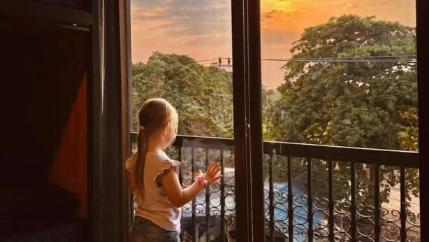 A child standing by a window with a view of trees and a sunset. - Friends Station Hostel & Cafe (Pattaya)