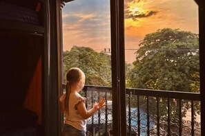 A child standing by a window with a view of trees and a sunset. - Friends Station Hostel & Cafe (Pattaya)