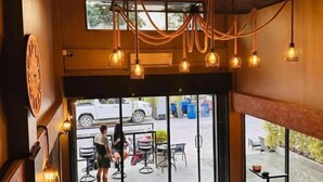 An interior view of a restaurant with hanging lights, a large window, and outdoor seating visible through the glass doors. - Friends Station Hostel & Cafe (Pattaya)