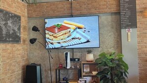 A brick wall with a mounted TV displaying a dessert, a small shelf with books and a plant, and a sign listing cities. - Friends Station Hostel & Cafe (Pattaya)