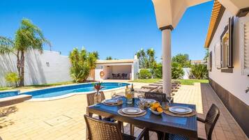 Un área de comedor junto a la piscina con una mesa preparada para cuatro, rodeada de sillas y una piscina al fondo.