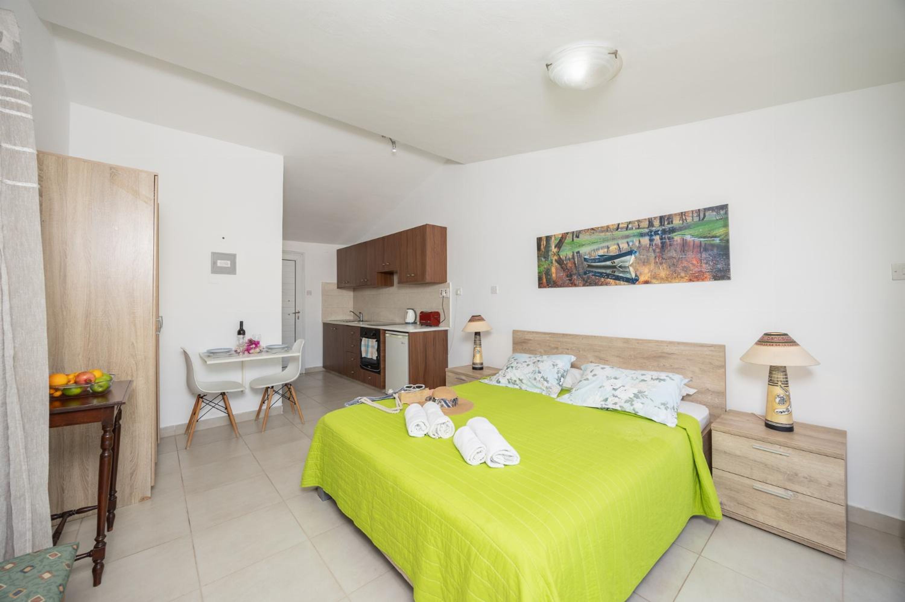 Habitación de hotel con cama, escritorio, silla, kitchenette pequeña y un cuadro en la pared.