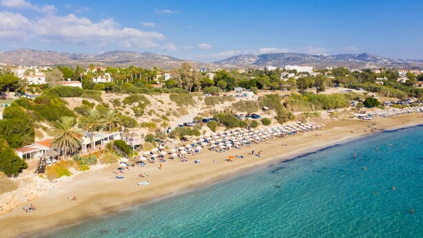 Una playa costera con aguas azules cristalinas, una orilla arenosa bordeada de sombrillas y edificios al fondo.