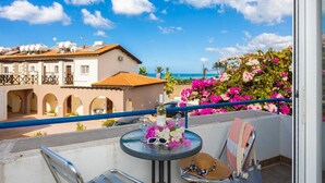Un balcón con una mesa para dos, con vistas a la costa, edificios y flores de buganvilla.
