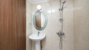 Un baño con puerta de madera, un lavamanos blanco de pedestal, un espejo redondo con marco texturizado y una ducha con múltiples cabezales.