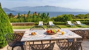 Un espace de restauration en plein air avec une table dressée pour deux personnes, entourée de chaises, offrant une vue panoramique sur la mer et les montagnes.