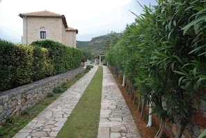 A stone pathway leading to a stone building with a tiled roof, surrounded by well-maintained hedges and trees. - Panorama II Villas in Ditiki Mani (West Mani)