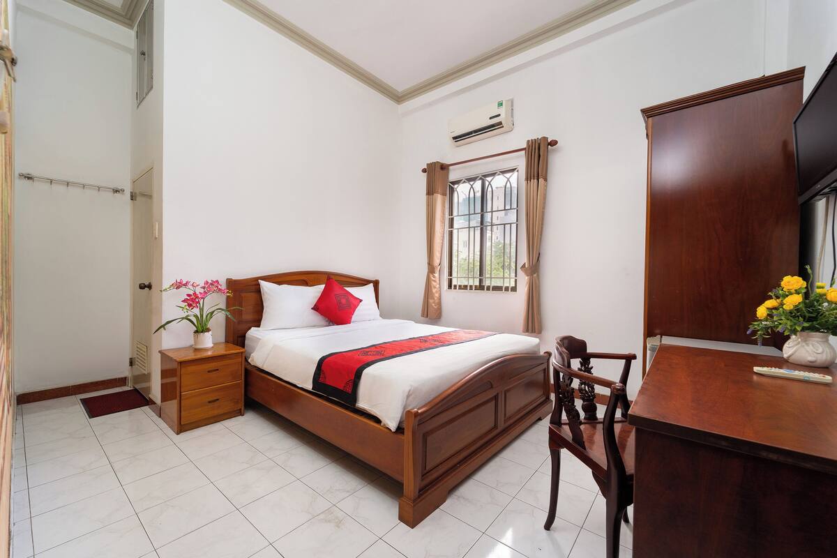 A neatly arranged bedroom with a wooden bed, a bedside table with a plant, a desk with a vase of flowers, and a window with curtains.