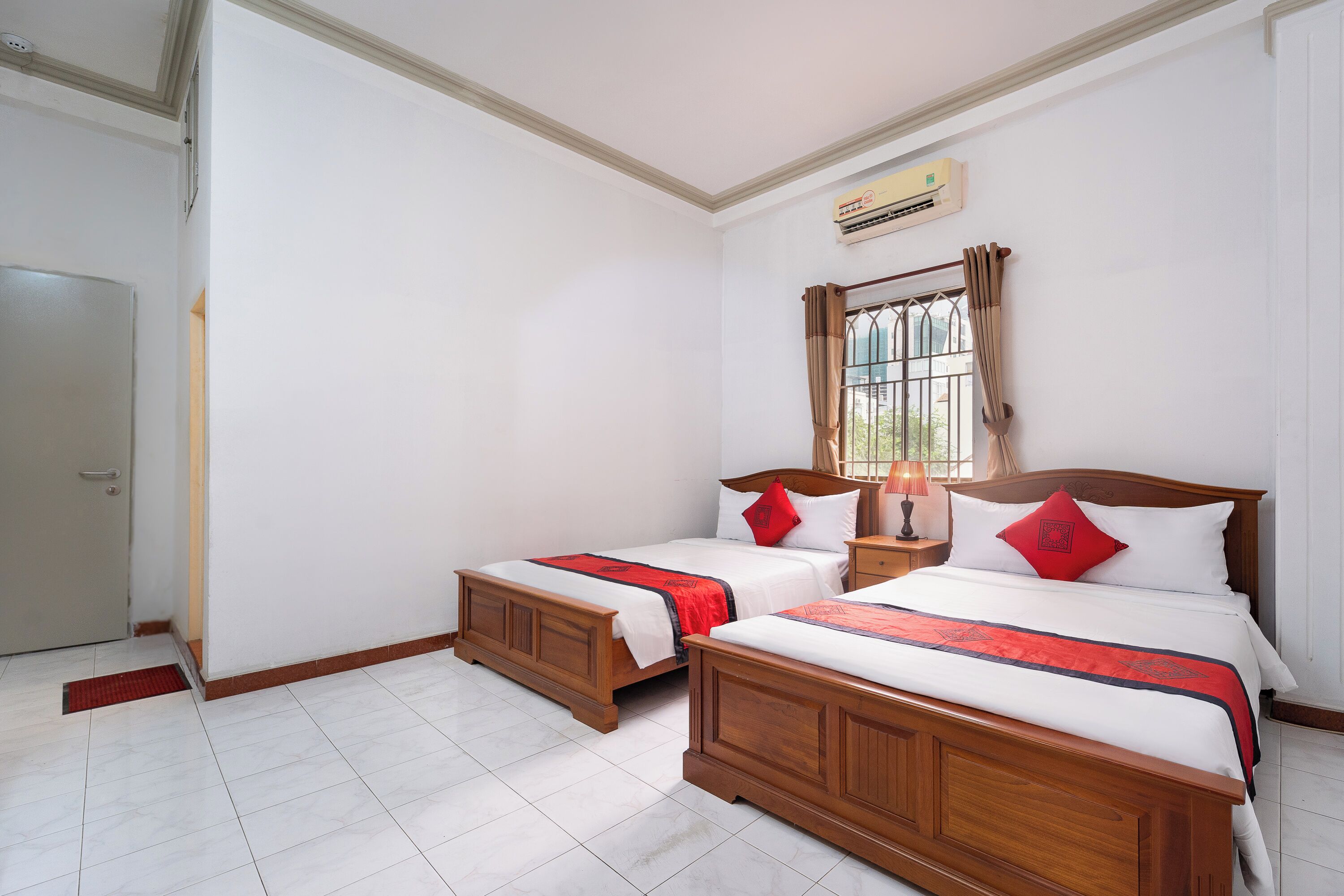 A hotel room with two wooden beds, a window with curtains, a bedside table with a lamp, and a wall-mounted air conditioner.