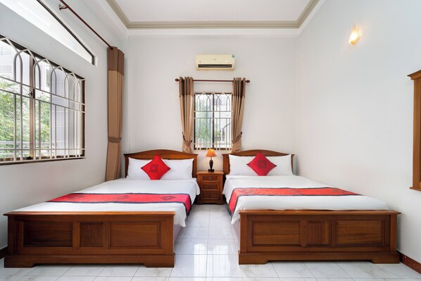 A hotel room with two wooden beds, a nightstand with a lamp, an air conditioner, and a window with curtains.
