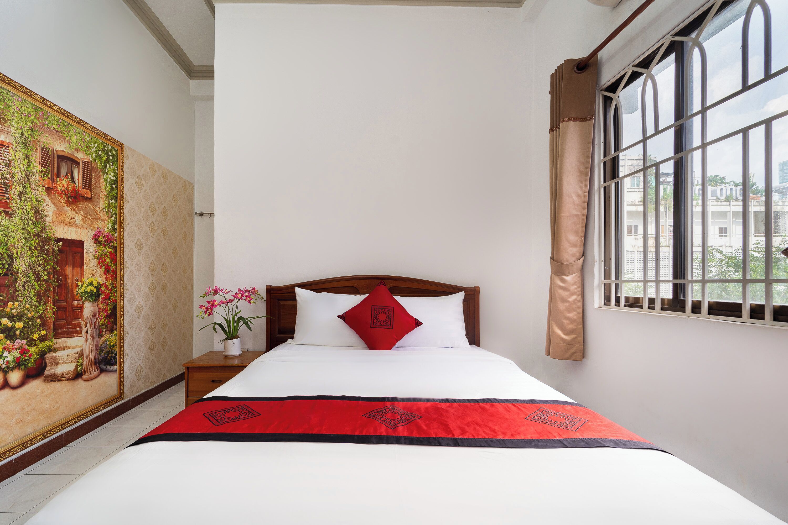 A neatly made bed with a red and black bedspread, white pillows, and a wooden headboard. A large floral painting adorns the wall to the left of the bed.
