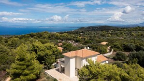 A white building with a tiled roof surrounded by dense greenery and a view of the sea. - Villa Eudora in Lefkada (Lefkada)