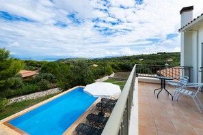 A swimming pool with a view of the countryside, a patio with chairs and a table, and a white umbrella.