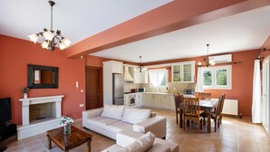 A living room with a sofa, a coffee table, a fireplace, and a kitchen area with cabinets and a dining table. - Villa Eudora in Lefkada (Lefkada)