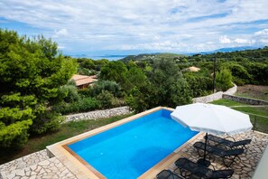 A rectangular swimming pool with a stone border, surrounded by greenery and a view of hills and the sea.