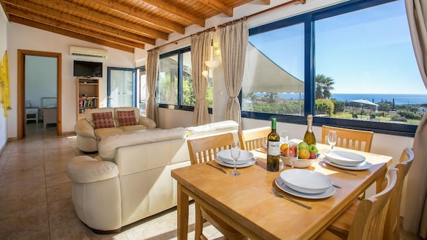 A spacious living area with a wooden ceiling, a sofa set, a dining table with plates and a bottle, and a large window offering a view of the landscape. - Villa Driftwood in Rhodes (Rhodes)