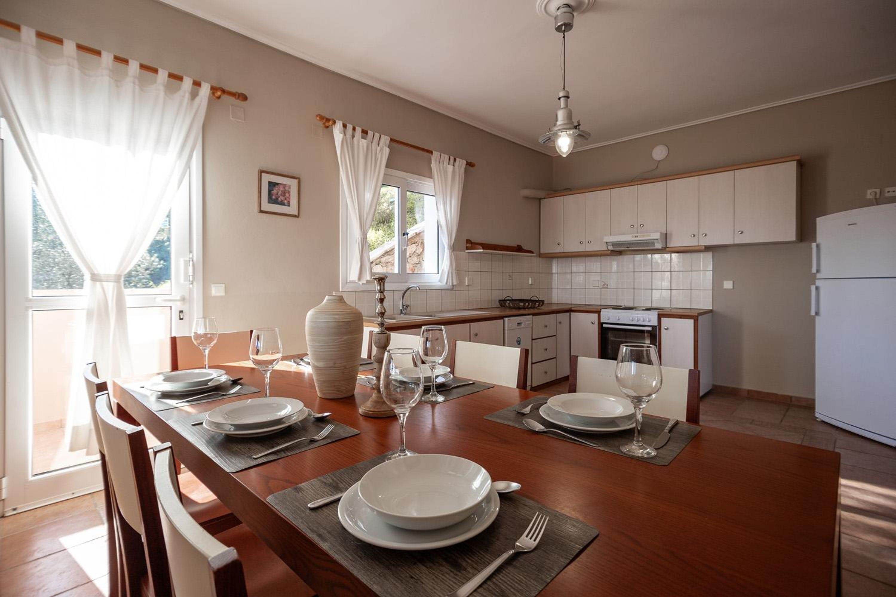 Un comedor con una mesa de madera dispuesta para cuatro personas, una cocina con muebles blancos y un ventanal con cortinas.