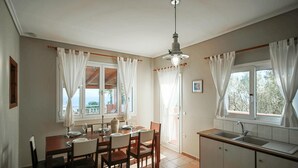 Ein Essbereich mit Tisch für vier Personen, eine Küche mit Spüle und große Fenster mit Vorhängen.